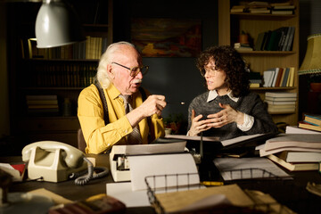 Senior Caucasian man discussing documents with young adult Caucasian woman at cluttered desk, both...