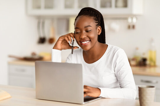 Telecommuting Concept. Portrait Of African American Woman Talking On Mobile Phone, Sitting At Desk And Working On Laptop, Cheerful Female Freelancer Using Pc And Having Pleasant Cellphone Conversation