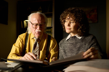 Senior Caucasian man explaining documents to young adult Caucasian woman at desk, both wearing...