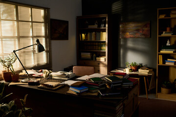 Office desk covered with stacks of books and papers standing in front of window with blinds,...