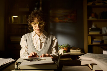Caucasian middle aged woman typing on typewriter in office surrounded by stacks of papers and...