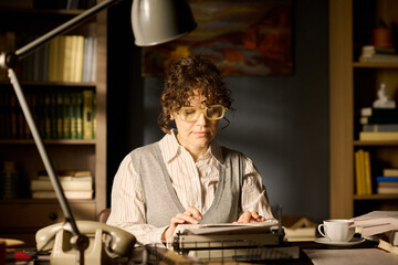 Caucasian middle aged woman typing on typewriter in office setting, wearing glasses and focusing on...