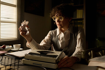 Caucasian middle aged woman sitting at desk holding crumpled paper in hand, wearing glasses,...
