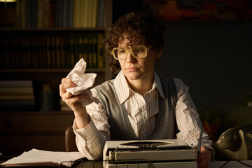 Caucasian middle aged woman sitting at desk holding crumpled paper in hand, looking at typewriter...