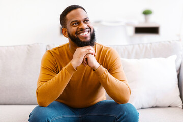 Dreamy Black Guy Thinking Smiling And Sitting On Couch At Home. African American Man Dreaming About...