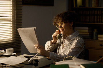 Middle aged Caucasian woman sitting at desk talking on landline phone while reading document, using...