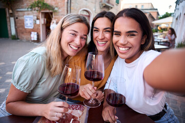 Three smiling multi-ethnic women are making a selfie while drinking red wine and eating a plate of cured meat outdoors in a restaurant in a small italian town