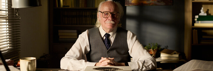 Portrait of senior Caucasian man sitting at desk with hands clasped, wearing glasses and formal...