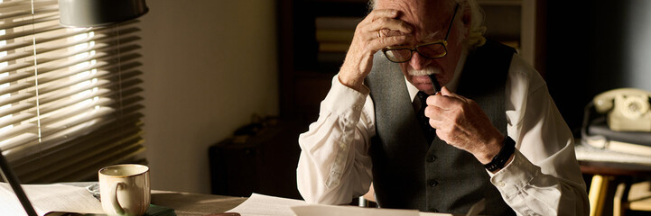 Senior Caucasian man sitting at desk reading documents, holding glasses and touching forehead,...
