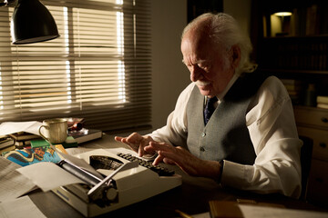 Senior Caucasian man sitting at desk typing on typewriter, concentrating on writing, surrounded by...