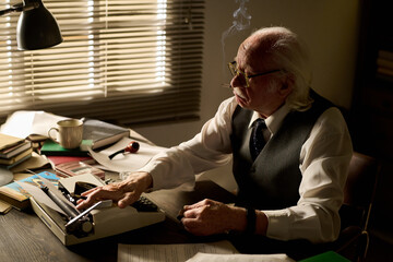 Senior Caucasian man wearing glasses sitting at desk typing on typewriter, surrounded by paperwork...