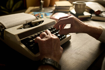 Senior Caucasian man typing on vintage typewriter with both hands, working at wooden desk with open book and coffee cup visible in background, focused on writing process