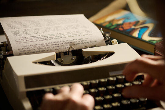 Caucasian adult hands typing on vintage typewriter with printed manuscript page visible, book with colorful cover lying nearby on wooden desk, focusing on creative writing process - Powered by Adobe