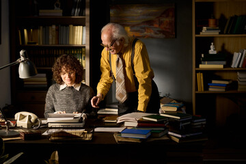 Middle aged Caucasian woman sitting at desk typing on typewriter while senior Caucasian man standing beside her pointing at paper, both surrounded by stacks of books in office setting