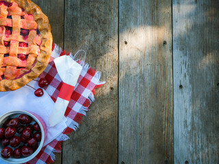 Overhead cherries and cherry pie on red checkered tablecloth on rustic weathered wood plank picnic country table with plastic silverware wrapped in napkin in dappled sunlight shadows
