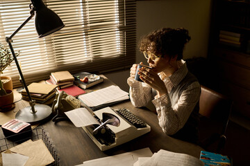 Young adult Caucasian woman sitting at desk drinking from mug while working with typewriter and...