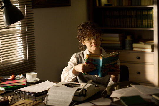 Middle aged Caucasian woman sitting at desk reading book, wearing glasses, surrounded by papers and typewriter, sunlight streaming through window blinds, bookshelf in background