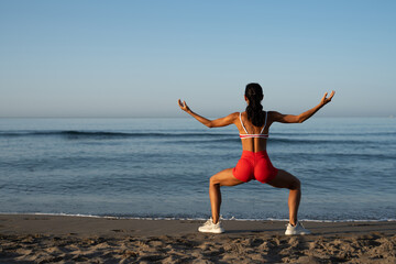 Athletic woman practicing grounding yoga pose on the beach at sunrise, facing the ocean with arms...