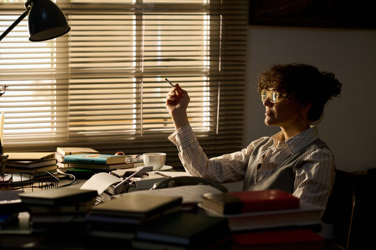 Caucasian young adult woman sitting at desk holding cigarette surrounded by stacks of books and papers, wearing glasses, gazing sideways with thoughtful expression