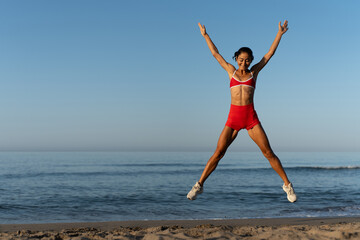 Energetic fit woman in red sportswear doing star jump on sandy beach at sunrise, showing strength,...