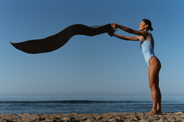 Young fit woman in blue swimsuit playfully shaking out a yoga mat on the beach at sunrise, smiling...