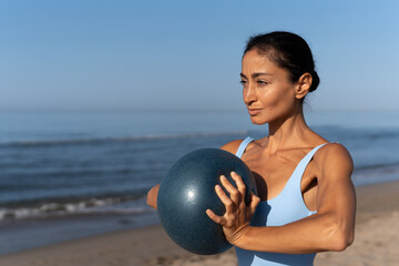 Focused athletic woman holding a fitness ball at chest level during a beach workout session at...