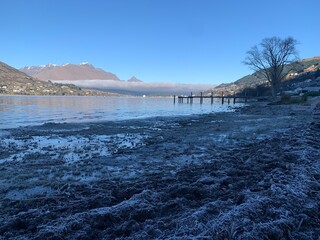 Frosty morning on lake surrounded by snow covered mountains, pier and dying leafless tree in the distance