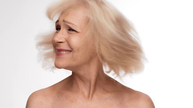 An elderly blonde woman in her 60s shaking her head while engaging in a pampering hair care routine. Set against a simple background, she radiates confidence and elegance. - Powered by Adobe
