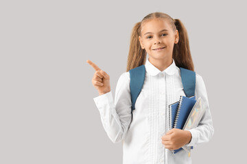 Cute little girl in school uniform with notebooks and pencil case pointing at something on grey...