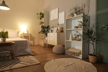 Interior of dark bedroom with cozy bed, houseplants and glowing lamp
