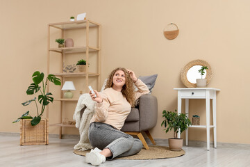 Beautiful smiling young woman with air conditioner remote control sitting on floor near armchair in...