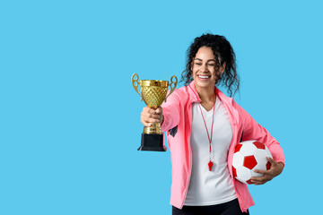 Female African-American soccer trainer with ball and gold cup on blue background