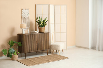 Interior of modern room with folding screen, pouf and chest of drawers near beige wall