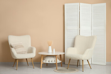 Interior of living room with armchairs, table and folding screen near beige wall