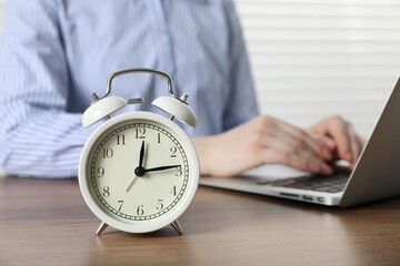 Deadline. Woman with laptop working at wooden table in office, focus on alarm clock