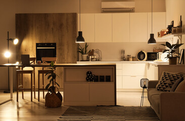 Interior of modern kitchen with counters, table and glowing lamps at night