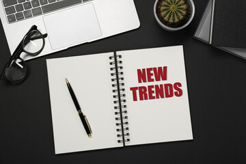 New Trends inscription in notebook, laptop, glasses and pen on black table, top view