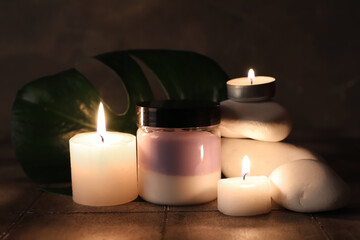 Jar of cosmetic product, spa stones and burning candles on grey tile background, closeup