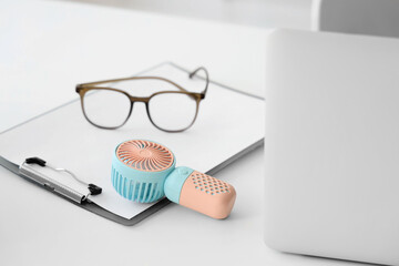 Small electric fan, clipboard, glasses and laptop on table in office