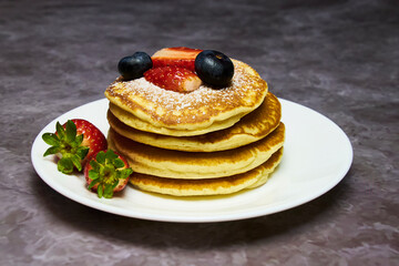 A plate of delicious pancakes in a tower with powdered sugar, decorated with blueberries and fresh berries (strawberries) for a wonderful breakfast.