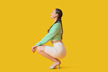 Stylish young woman with dreadlocks sitting on yellow background