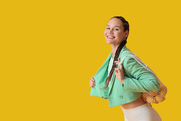 Stylish young woman with dreadlocks and string bag on yellow background