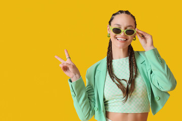 Stylish young woman with dreadlocks and sunglasses showing victory gesture on yellow background
