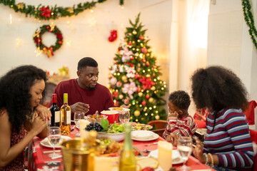 A cheerful holiday dinner scene with a diverse family enjoying a festive meal. Christmas decorations, gifts, and a lit Christmas tree create a cozy atmosphere, enhancing the warmth of the celebration.