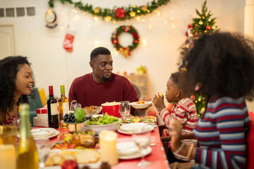 A warm Christmas family dinner with a father serving food to his young child, surrounded by festive decorations, a Christmas tree, and a holiday table spread. Joyful and cozy holiday atmosphere.