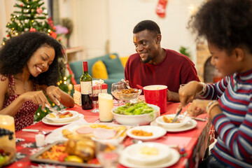 A joyful family shares a Christmas meal together, passing food and creating holiday memories. Decorated tree, festive lights, and presents in the background add warmth to cozy celebration atmosphere.