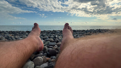 Horizontal travel wide angle photo. Sun day. Male hairy bare legs shoes close up on Black Sea pebble beach. Blue cloudy sky. Ocean waves. Concept of coast line, rest, summer vacation, tourism