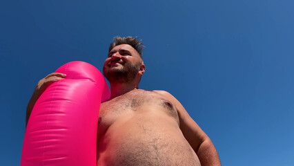 Horizontal mobile photo. Bottom view. Fat mature white man with beard, big belly, pink swim ring on sea beach put hand to forehead to shield eyes from sun shine. Sun day. Blue sky. Travel, trip	