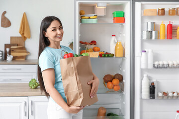 Beautiful woman with paper bag of food near open fridge in kitchen