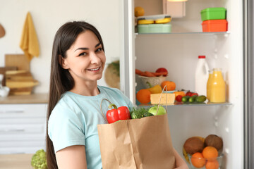 Beautiful woman with paper bag of food near open fridge in kitchen